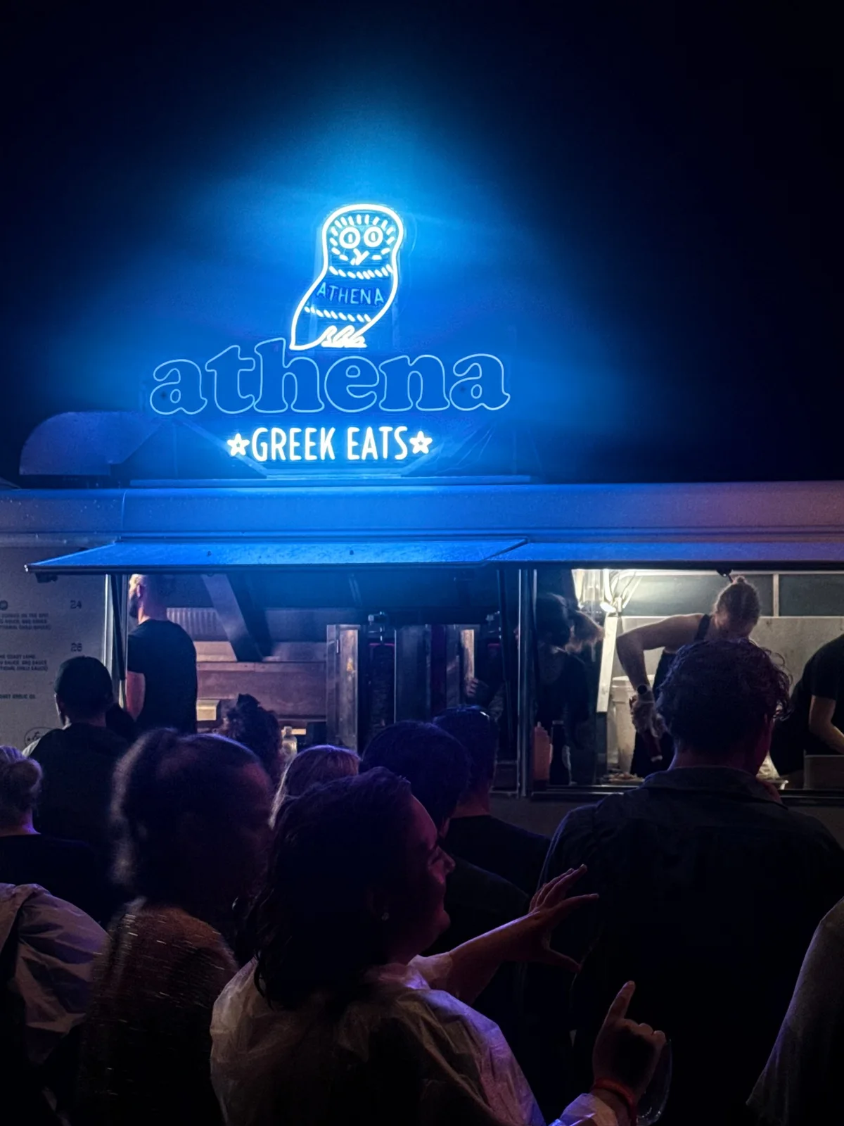 A crowd ordering from the Athena Greek Eats truck beneath its glowing blue owl sign at night
